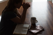 a woman sitting at a table praying and reading a Bible 