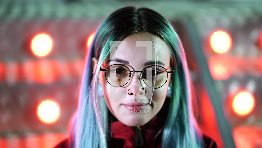 Hipster girl with blue dyed hair, golden sequins as freckles. Woman with nose piercing, transparent glasses, ears tunnels, unusual hairstyle stands in amusement night park