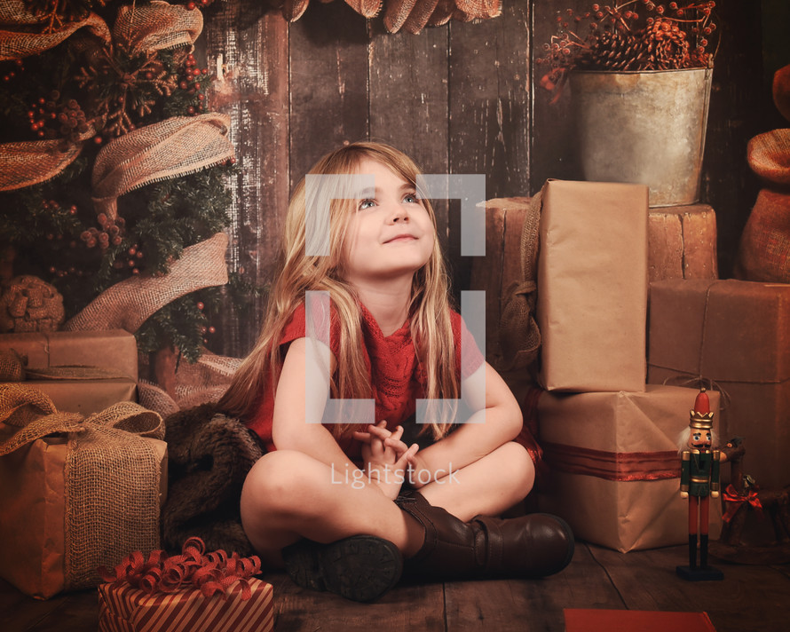 a little girl sitting next to Christmas presents with praying hands 