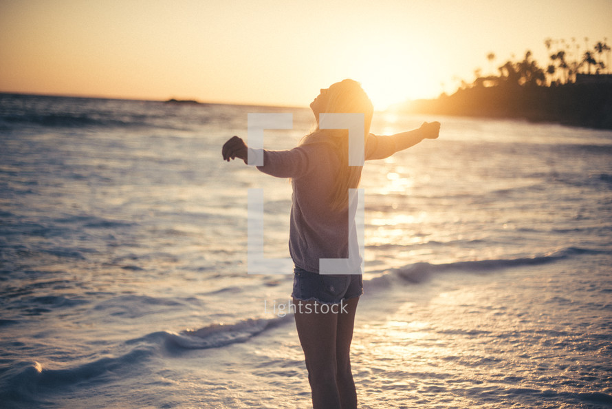 a woman with outstretched arms standing on a beach at sunset 