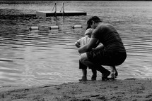 father and son squatting at the edge of a lake 