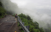 Two men with a motorcycle on the side of a winding mountain road.