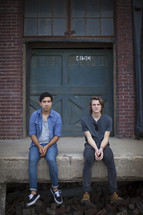 Two men sitting on a stoop in front of a warehouse 