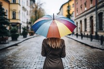 A girl walking in the rain through a gray city, carrying a rainbow umbrella. The view is from behind