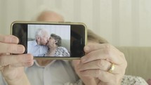 Senior caucasian couple holding up a smartphone to the camera with an image of themselves kissing then revealing themselves behind themes of love romance technology