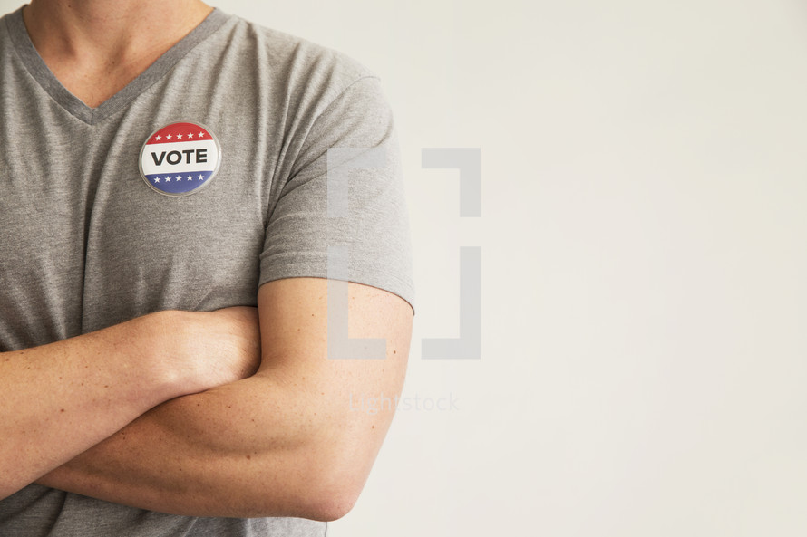 A man with arms crossed wearing a red white and blue button reading, "Vote."