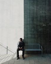 A man sitting on a metal bench against a tall concrete wall.