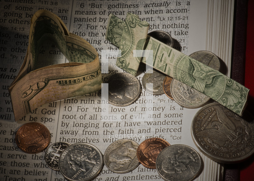 Paper money heart and cross with coins on pages of Bible open to 1 Timothy 6:10 -- "for the love of money."