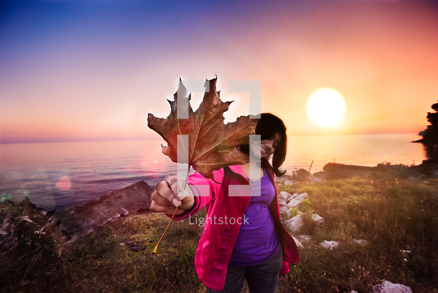 girl holding up a large dead leaf in front of the sun at sunrise