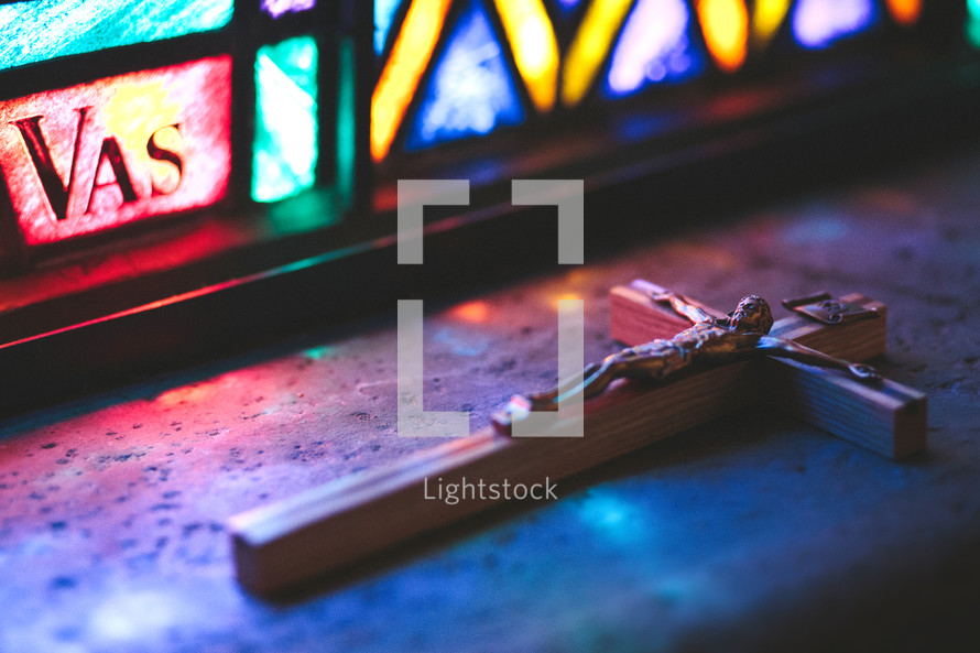 Crucifix sitting in colored light on ledge near stained glass in a Roman Catholic church.