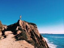 Man standing on a rocky cliff at the beach overlooking the ocean.