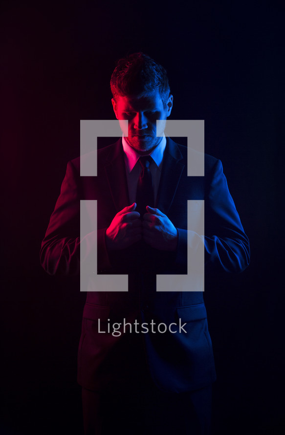 Red and blue light shining on a man who is looking down and holding the lapel of his coat.