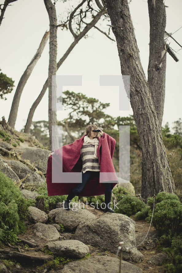 Woman in a red cape looking through binoculars, standing on a rock in a tree-filled forest.