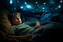A young boy peacefully rests in bed, enveloped by the gentle, soft blue glow of star projections