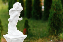 statue of baby angel with wings against green garden. The cute little cupid angel sculpture statue standing outdoor.