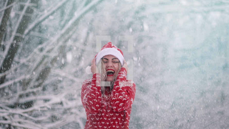 Cheerful woman enjoys winter wonderland. Snow flies on her and she is happy, rejoices cold weather. Girl in red Christmas sweater and Santa hat. High quality photo