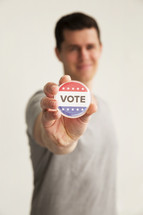 A man holding out a red white and blue button reading, "Vote."