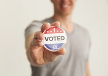 A smiling man holding out a red white and blue button reading, "I Voted."
