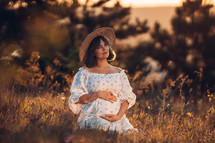 Happy Mother Carrying Child, Expecting For Baby. Amazing Motherhood. Pregnant Woman in beautiful dress and straw hat sitting on grass at nature rural meadow. High quality 