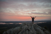 Silhouette of a person with arms raised standing on a mountain at sunrise.