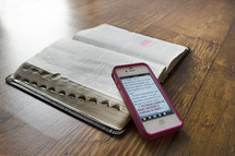 cellphone on the pages of an open Bible on a wood floor 