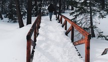 man walking across a snow covered bridge into a forest 