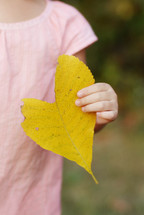 A little girl in a pink dress holding a bright yellow leaf.