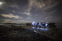 "Grace" in neon lights on rocks in the ocean water.