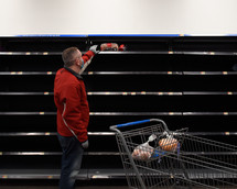 A man is wearing a medical mask at a grocery store reaching for bread with empty shelves for a pandemic emergency concept.