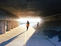 A woman and child walking under a concrete  bridge.