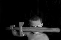 a boy child holding a wooden sword  across his face with a black background