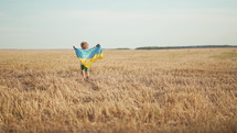 Happy little boy - Ukrainian patriot child running with national flag in field after collection wheat, open area. Ukraine, peace, independence, freedom, win in war.