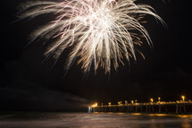Fireworks exploding over a pier extending into the ocean.