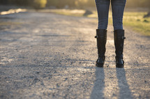 a woman in knee high boots standing on a dirt road 