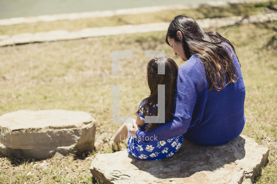 Mother embracing daughter while sitting on a rock in a park.