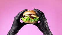 Hands in black latex gloves with burger. Woman holding hamburger. Big appetizing patty with meat cutlet, vegetables, cheese. Fast food concept. Pink background. High quality photo