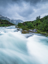 A river surrounded by lush forest, cascading over rocky rapids and framed by an ethereal sky of soft clouds. A tranquil beauty in the heart of Norway.