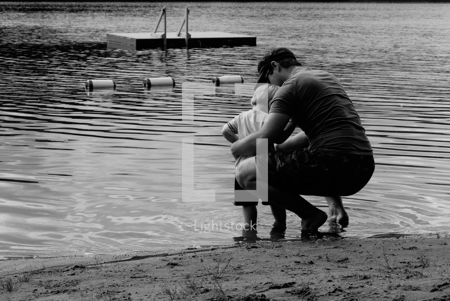 father and son squatting at the edge of a lake 