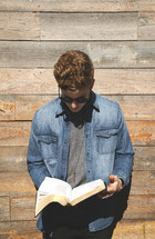 a young man reading a Bible in front of a wall of wood boards 