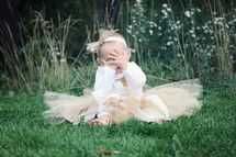 an infant girl in a tutu sitting in the grass covering her face 