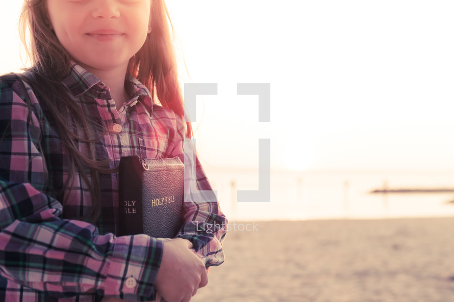 young girl happy, smiling, holding holy bible, standing on the beach at sunset