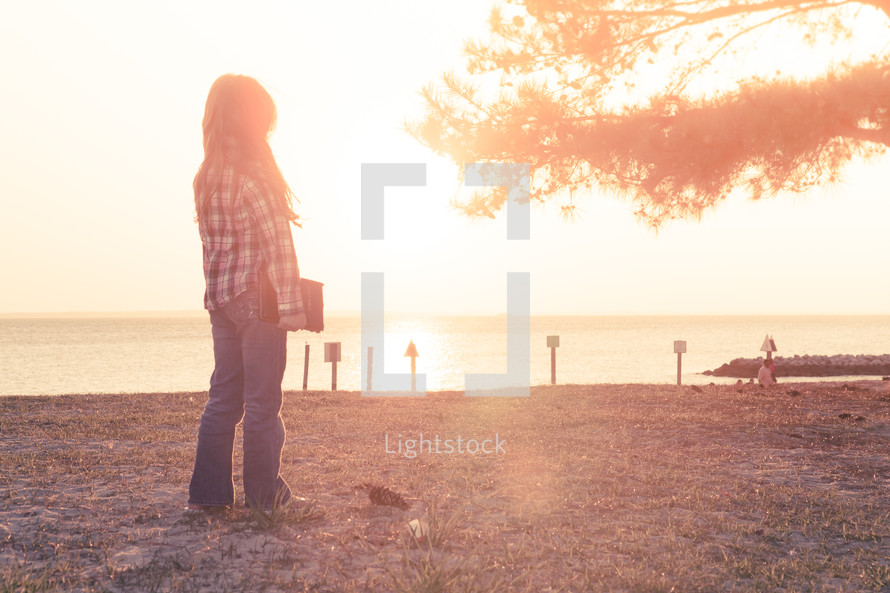 young girl standing on the beach holding her bible at sunset; thinking, nature, sunflare