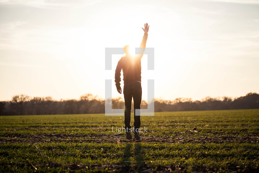 silhouette of a man jumping in a field with his hand raised to God glowing under sunlight