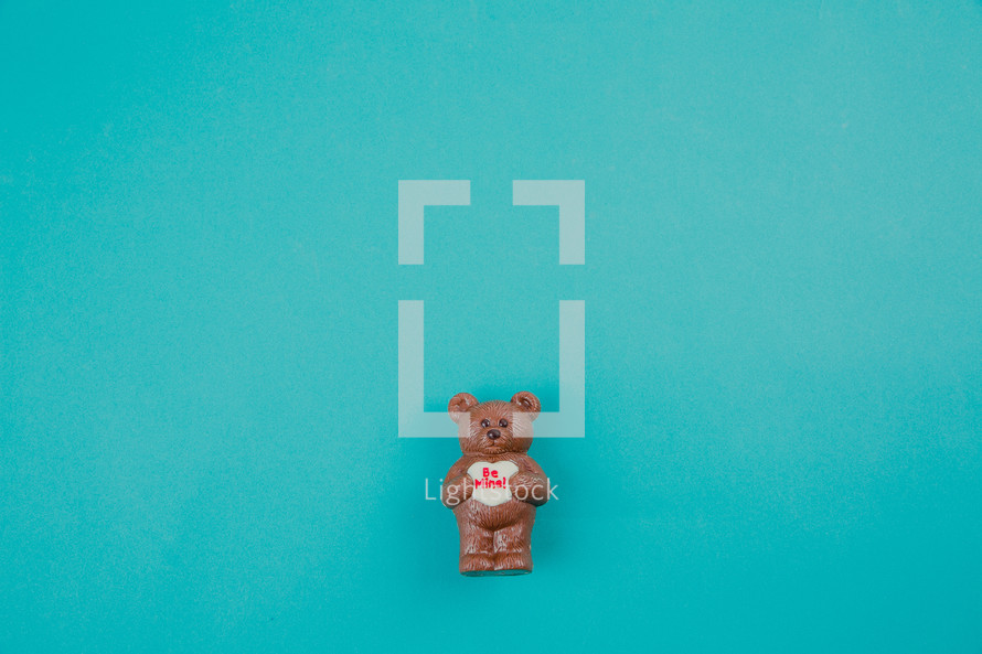 A chocolate bear holding a heart that says "be mine" on an aqua background.