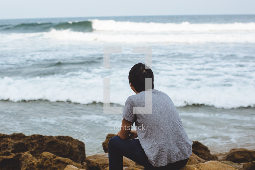 A man sitting on a rocky shore looking out at the ocean.