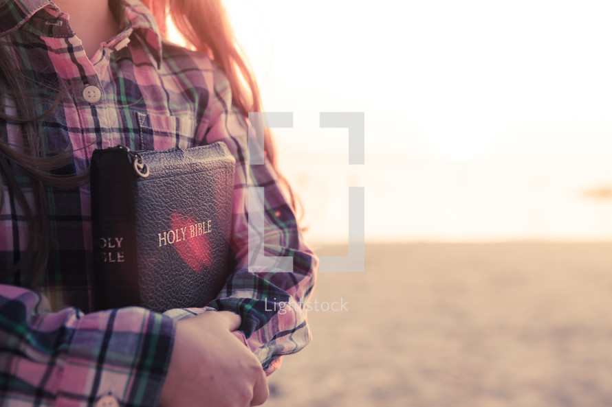 young girl holding holy bible, standing on the beach at sunset