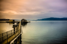 Pier leading to a boathouse on the water with mountains on the horizon at dusk.