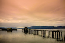 Pier leading to a boathouse in the water with mountains on the horizon at dusk.