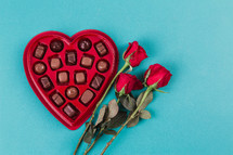 A heart shaped box of chocolates and three red roses.