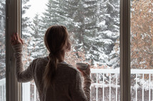 a woman standing in a window watching falling snow 